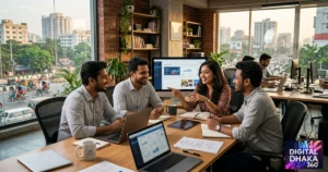 A group of professionals in the Digital Dhaka 360 office is discussing at a wooden table with laptops.